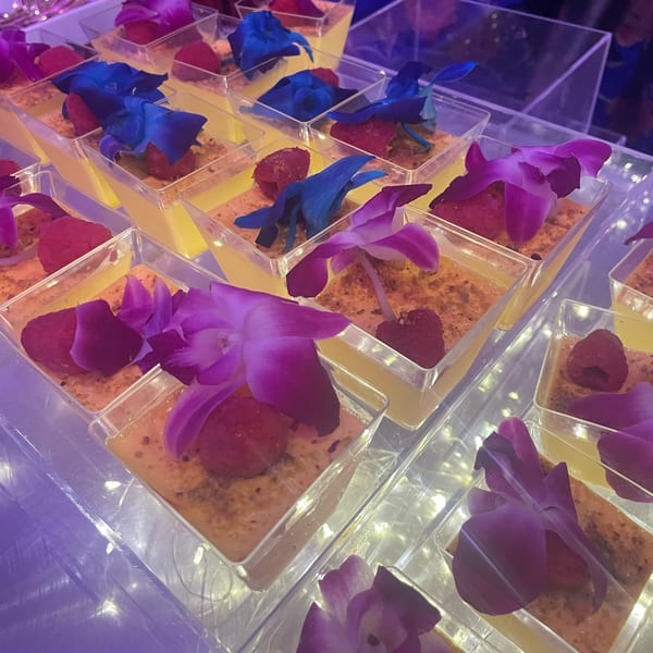 Rows of individual desserts in clear cups, garnished with raspberries and colorful orchid petals.