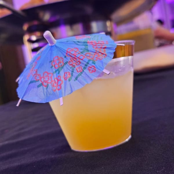 A vibrant yellow cocktail with ice and a blue paper umbrella sits on a dark surface.
