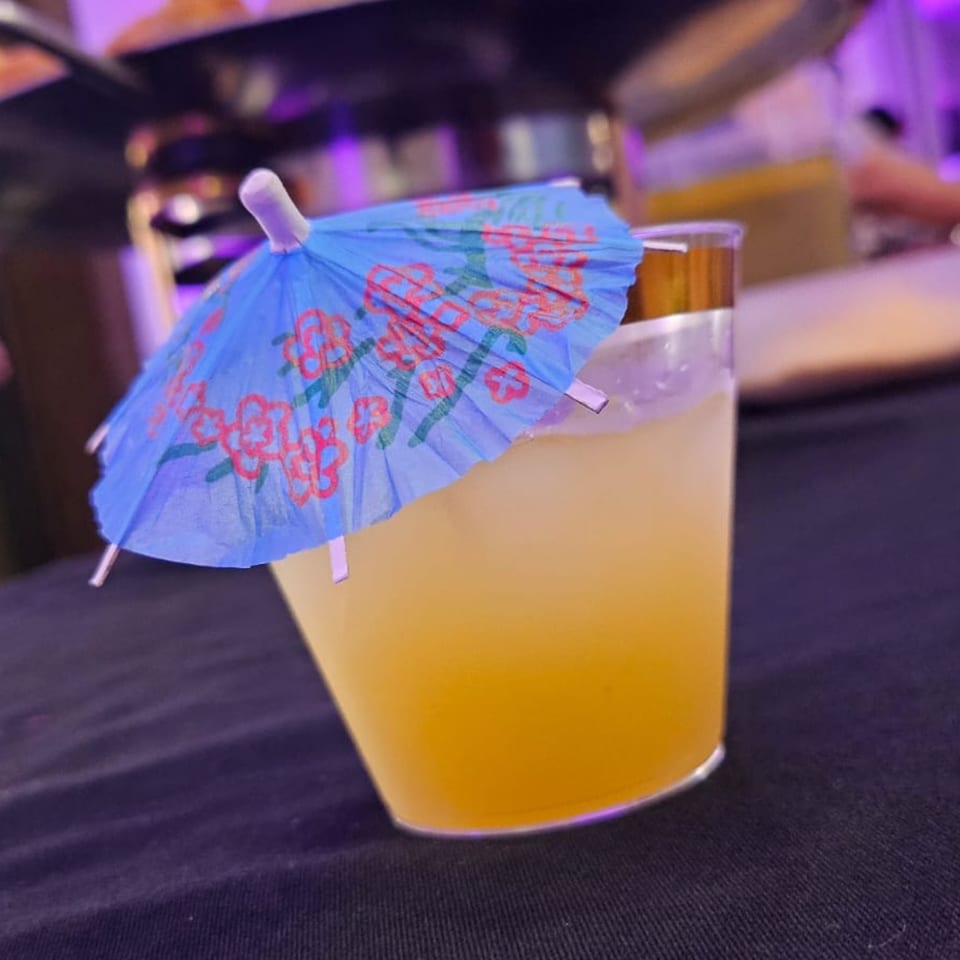 A vibrant yellow cocktail with ice and a blue paper umbrella sits on a dark surface.