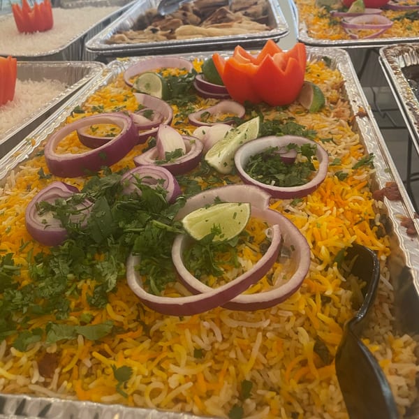 Biryani rice in a foil tray, garnished with red onions, cilantro, lime, and a carved tomato.