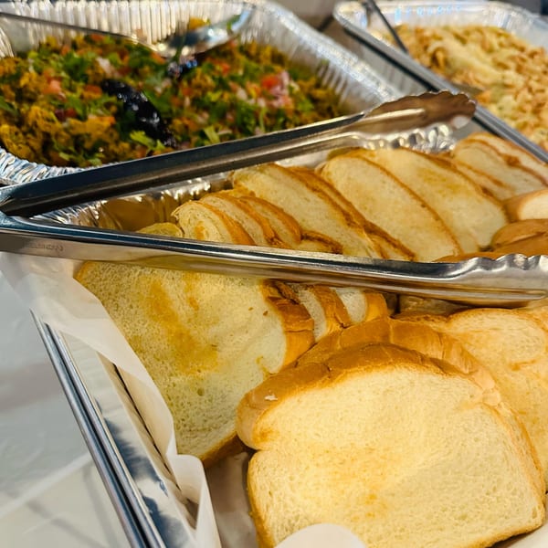 Close-up of sliced bread in a serving tray with tongs, and a blurred dish of food in the background.