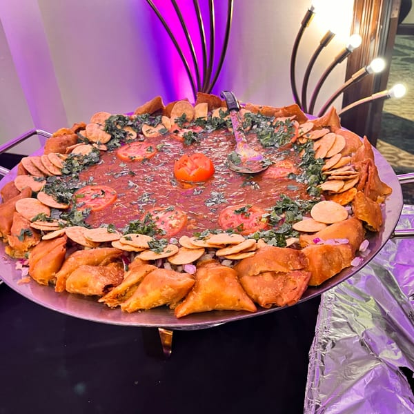 Large platter of Indian chaat with samosas, papdi, tomatoes, and cilantro, under purple lighting.