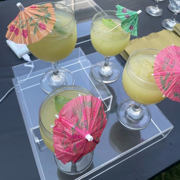 Four cocktails with colorful paper umbrellas and mint garnishes on clear acrylic trays.